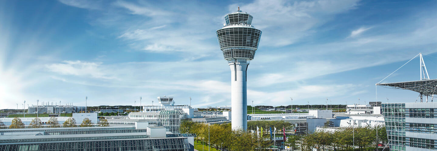 Net Zero by 2035 View of Munich Airport's tower, flanked by the characteristic wave-shaped terminal roofs under a bright blue sky. The scene appears modern, spacious, and technically impressive.