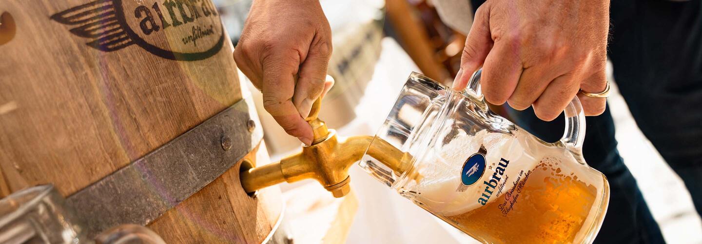 Host and Brewer Two people pour fresh beer from a traditional wooden barrel into an Airbräu mug. The scene has a down-to-earth, Bavarian, and festive feel—typical beer culture at Munich Airport.