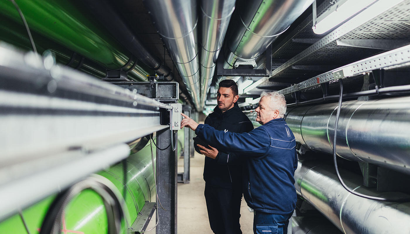 Technik - erneuerbare Energien, Ver- und Entsorgung Zwei Personen inspizieren technische Anlagen in einem unterirdischen Versorgungsgang mit großen Metallrohren und grünen Leitungen.