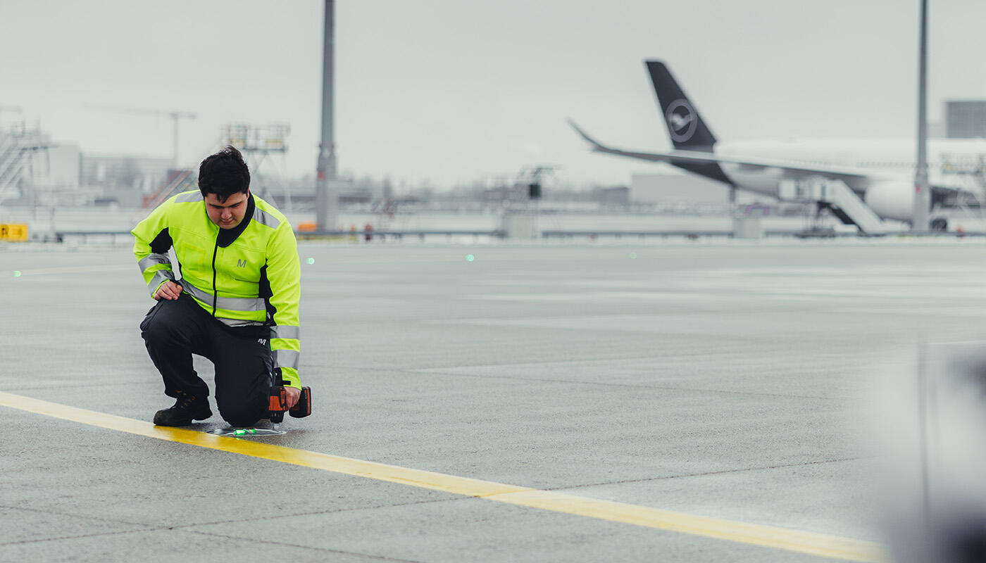 Technik - Luftverkehrsflächen und -Anlagen Person in Warnjacke überprüft ein Licht auf dem Rollfeld eines Flughafens, im Hintergrund steht ein Flugzeug.