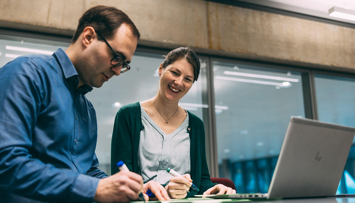 Technik - kaufmännische Steuerung Zwei Personen arbeiten gemeinsam an einem Tisch mit Laptop und Dokumenten in einem modernen Büro, beide halten Stifte und machen Notizen.