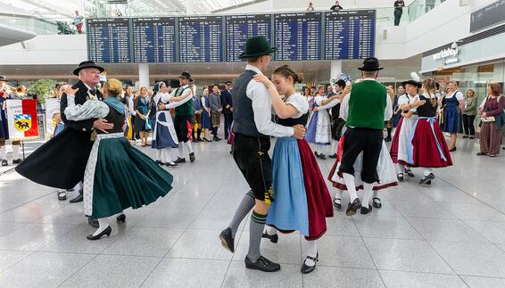Oktoberfest 2026 Group of people in traditional Bavarian dress dancing in an airport terminal, with a large departure board and waiting travelers in the background.